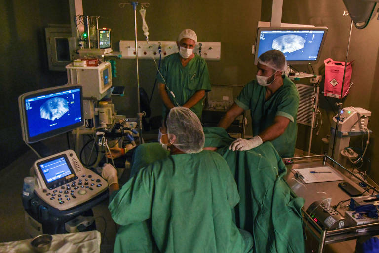 Três profissionais de saúde em trajes cirúrgicos concentram-se em um procedimento, com um deles operando um equipamento de ultrassom. Monitores ao fundo exibem imagens médicas, iluminando a sala escura com tons de azul e verde.