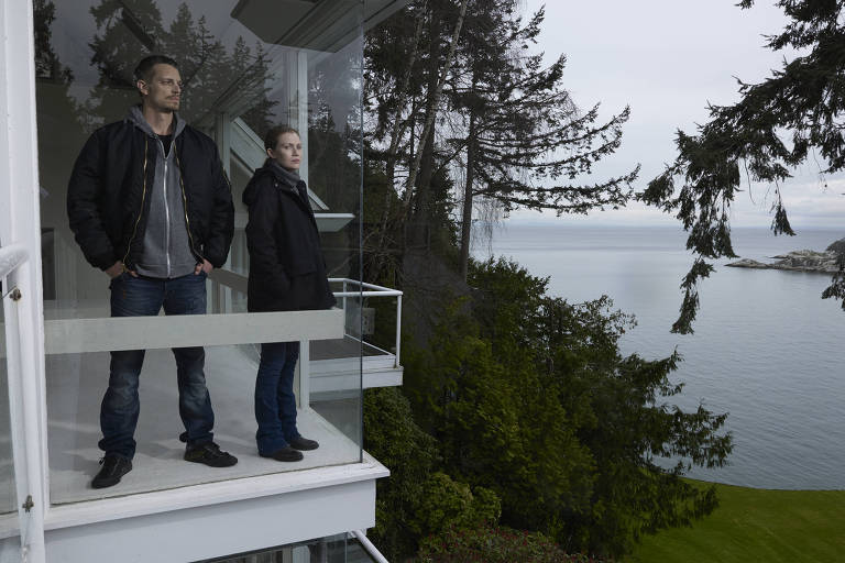 Um homem e uma mulher, vestidos de roupas de frio de tons escuros, estão em uma varanda fechada com vidros em uma casa ao lado de um lago. Eles têm expressões sérias