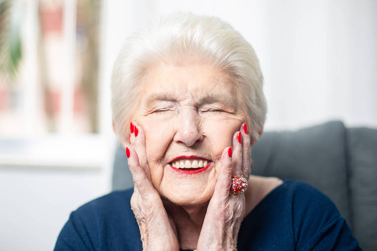 Estelinha Bezerra, de 94 anos, posa para a foto com os olhos fechados, sorridente e com as mãos nas bochecas; ela tem cabelos brancos curtos, unhas e batom vermelhos e veste uma blusa azul