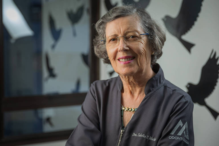 Zuleika Machado, de 80 anos, veste uniforme cinza com seu nome bordado e posa para a foto com um sorriso; ela usa óculos de grau, tem cabelos grisalhos e, ao fundo, há adesivos de pássaros colados na parede