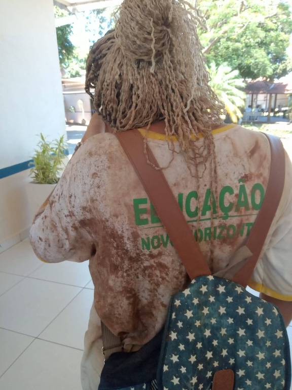 foto de adolescente negra de costas, com tranças loiras, e uniforme escolar escrito Educação Novo Horizonte sujo de terra, com mochila