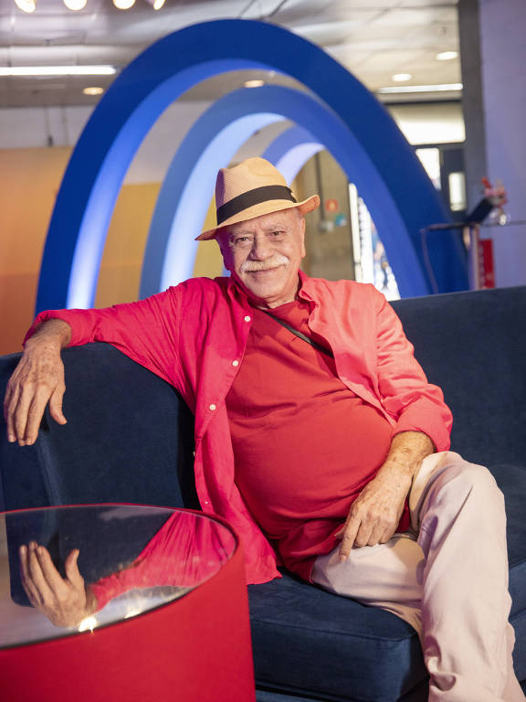 Ator Tonico Pereira está sentado em sofá; ele veste chapéu bege, camiseta e camisa vermelhas e calça bege. Está com as pernas cruzadas e um dos braços apoiados sobre o sofá