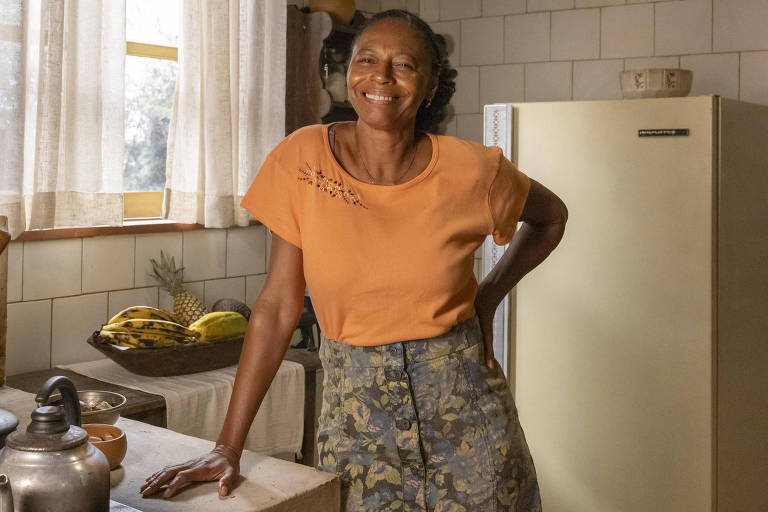 Em foto colorida, mulher sorri na cozinha de um cenário de novela