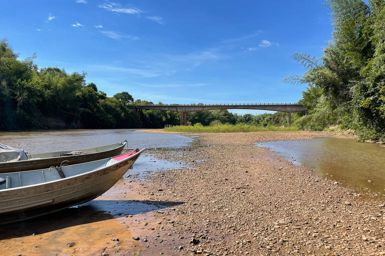 Barco em trecho seco do rio; chão está sem água, apenas coberto de cascalho; ao fundo há uma ponte