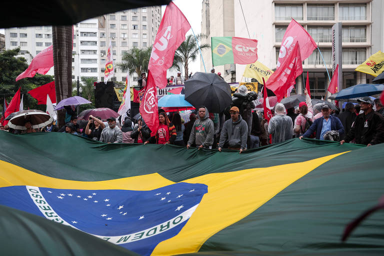 Imagem contém vários manifestantes segurando a bandeira do Brasil, de cores verde, amarela, azul e branca, e outros segurando bandeiras vermelhas de movimentos sociais