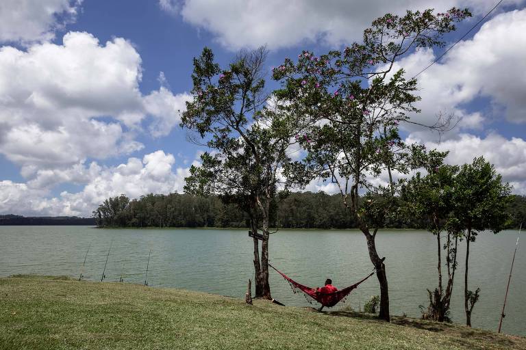 Imagem geral mostra uma pessoa sentada na rede presa entre duas árvores, em ambiente bucólico. A frente é possível ver um lago e vegeteção ao longe. Céu azul com nuvens.