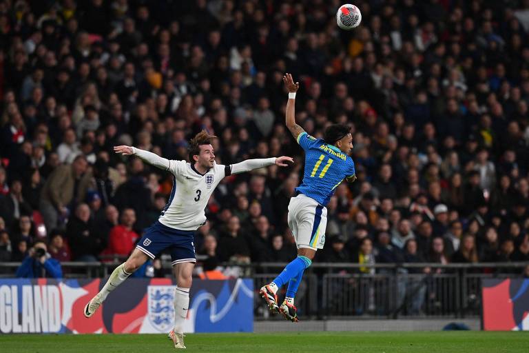 Os dois jogadores estão saltando no ar, com a bola muito acima dos dois. O inglês tem os braços abertos, e o brasileiro, o braço esquerdo acima da cabeça