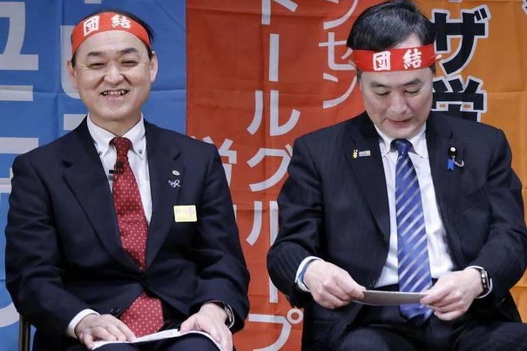 Imagem mostra dois homens sentados lado a lado. Eles usam terno e uma faixa na cabeça.