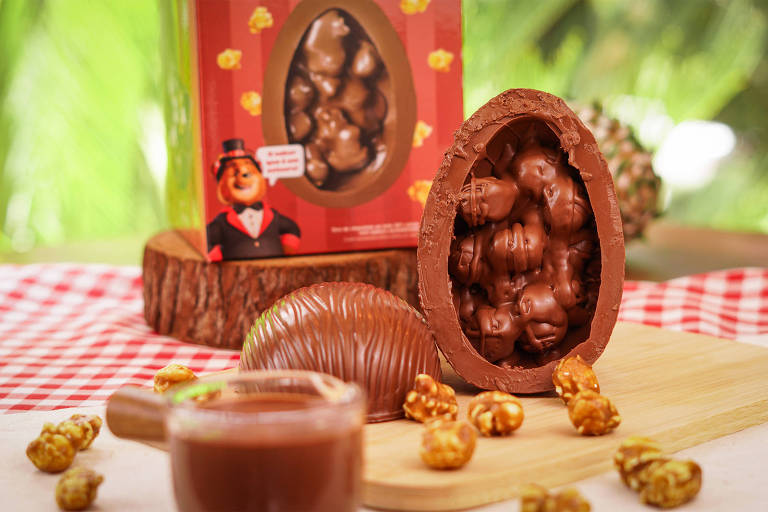 A foto mostra uma casca de ovo de Páscoa recheado de pipoca caramelizada coberta com chocolate. Ao fundo uma caixa vermelha com uma foto do produto