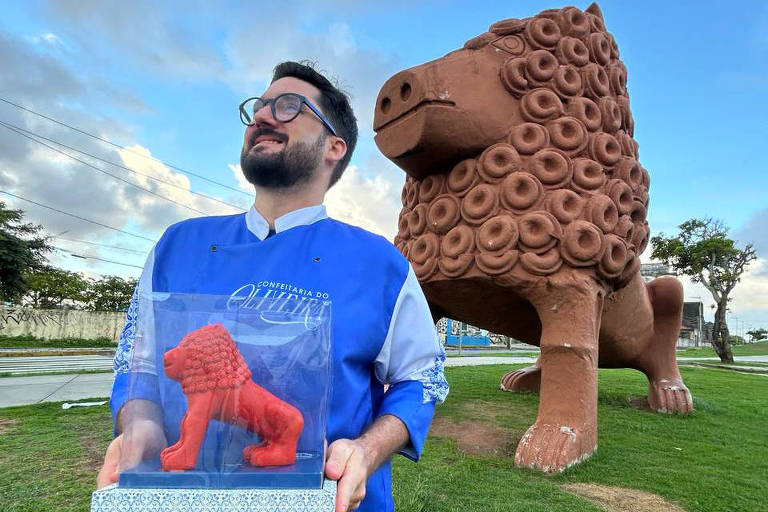 A foto mostra um homem segurando uma caixa com uma escultura de uma reprodução do Leão de Nuca feita com chocolate. Ao fundo, a escultura feita em cerâmica. 
