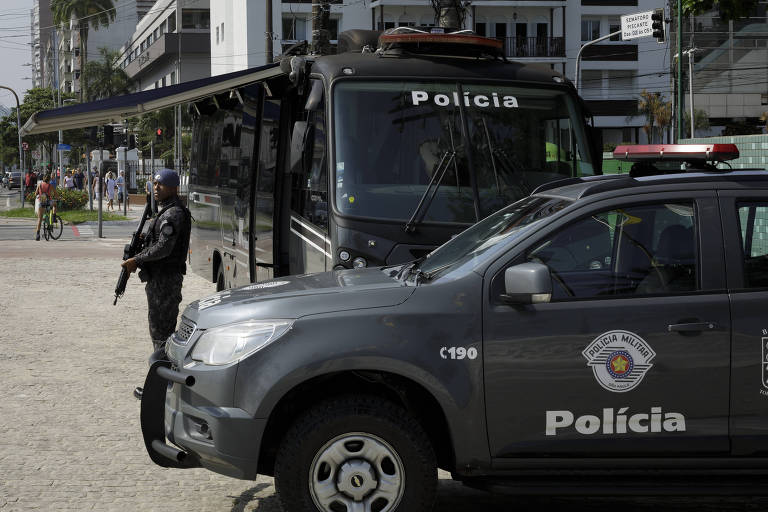 Um policial armado em pé ao lado de um carro policial. Ao fundo， há um ônibus da polícia estacionado， com a palavra 039;Polícia039; visível. O ambiente é urbano， com prédios e árvores ao redor.