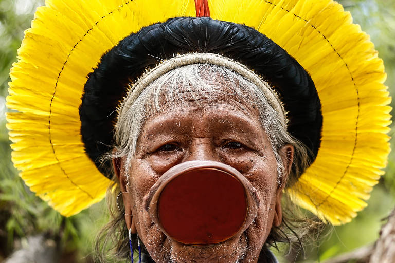 Usando cocar de penas amarelas e pretas e um grande disco de madeira, o bodoque, nos lábios, idoso indígena olha para a câmera