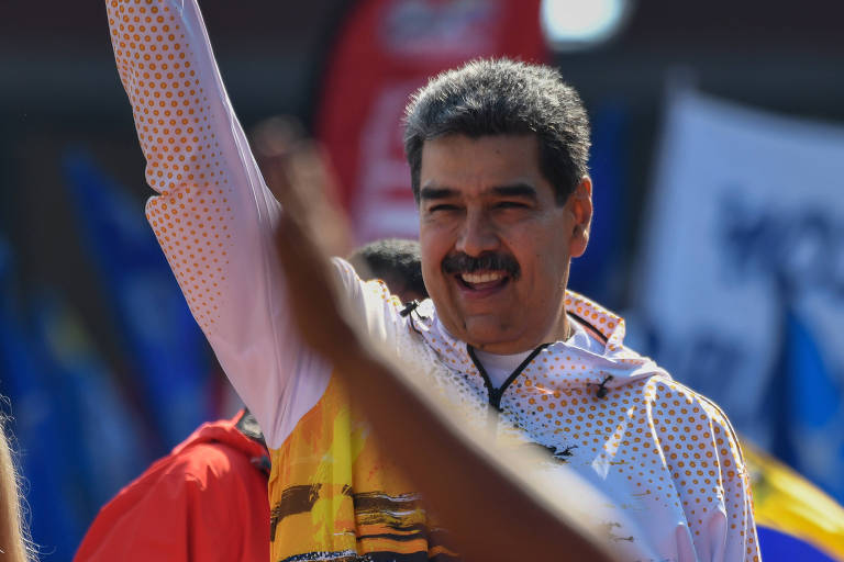 O ditadir venezuelano Nicolás Maduro, um homem branco, de cabelo e bigode pretos, ergue o braço direito, enquanto podemos ver o braço de outra pessoa levantado também à sua frente