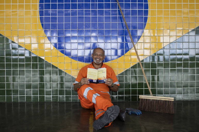 gari com roupas laranja segura livro ao lado de vassoura, com painel de bandeira do Brasil em azulejo ao fundo