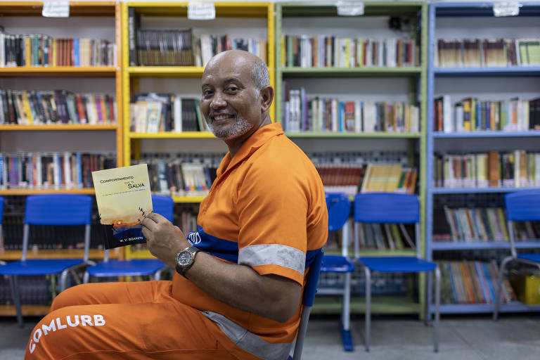 homem com roupa de varredor de rua laranja escrito Comlurb segura livro sentado lateralmente com livros ao fundo