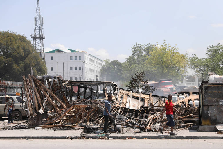 Uma série de escobros de carros queimados são vistas em rua; em frente, uma pessoa caminha; ao fundo, vê-se o palácio presidencial
