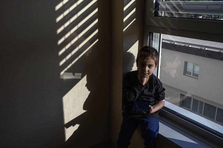 Garoto branco de cinco anos está perto de uma janela, que tem persiana refletida na parede com a luz solar