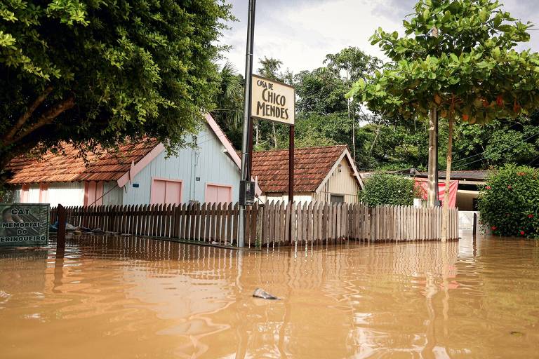 Casa inundadas pela água após enchente que atingiu o município de Xapuri, no interior do Acre. A imagem mostra duas residências cercadas por muros de madeira, uma delas tem uma placa escrito Casa de Chico Mendes