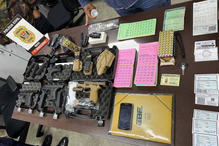 cartelas de rifa, armas e munições sobre mesa de madeira, com o brasão da polícia civil ao lado esquerdo