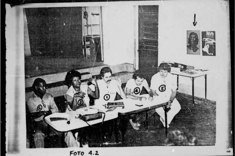 Uma sala de aula, com cinco pessoas em uma mesa, quatro homens e uma mulher negra. Na parede, uma foto de Che Guevara