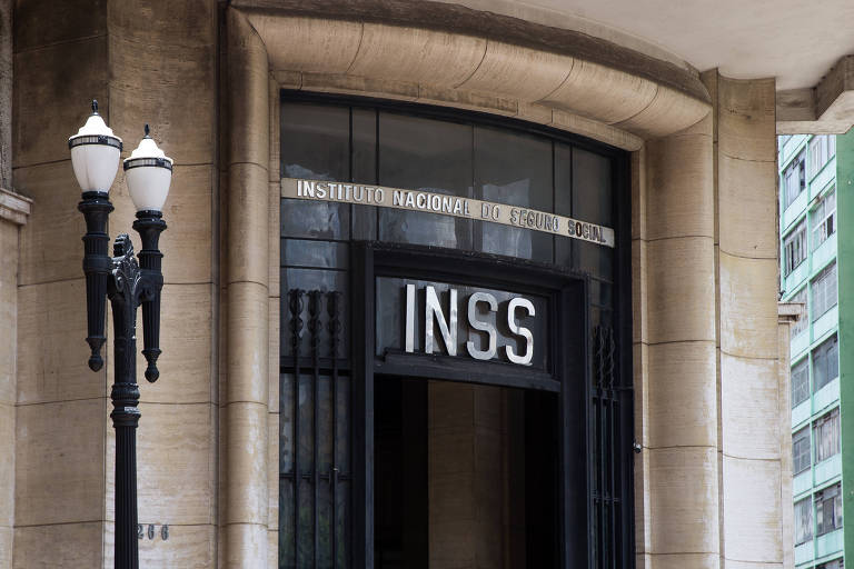 Imagem da entrada de um prédio com a inscrição 'INSS' acima da porta. A fachada é de pedra e há uma luminária de rua com dois postes ao lado da entrada. O prédio tem um design arquitetônico clássico.