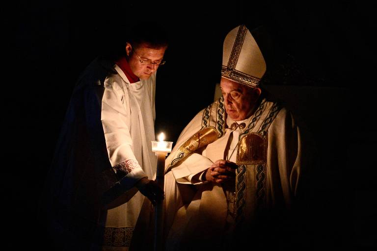 À direita, o Papa Francisco, vestido com manto, olha para uma vela em um ambiente escuro