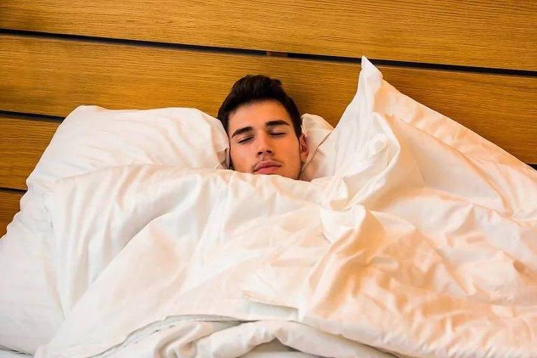 Homem branco dormindo numa cama com lençóis brancos