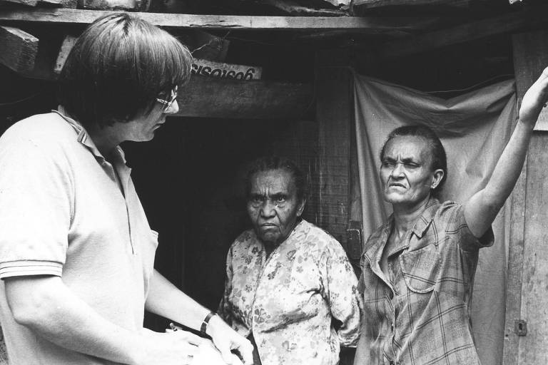 Pesquisador do IBGE entrevistando duas mulheres na porta de uma casa simples. O IBGE enviou 1200 pesquisadores para investigar o consumo familiar em 1974 e 1975