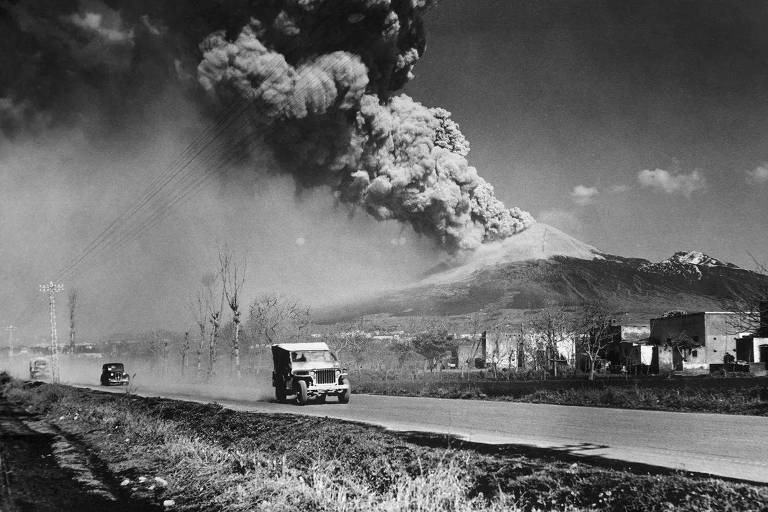 Carros passando em estrada com o Vesúvio em erupção ao fundo. A erupção de 1944 aconteceu durante a Segunda Guerra Mundial, mas agora está desaparecendo do imaginário coletivo