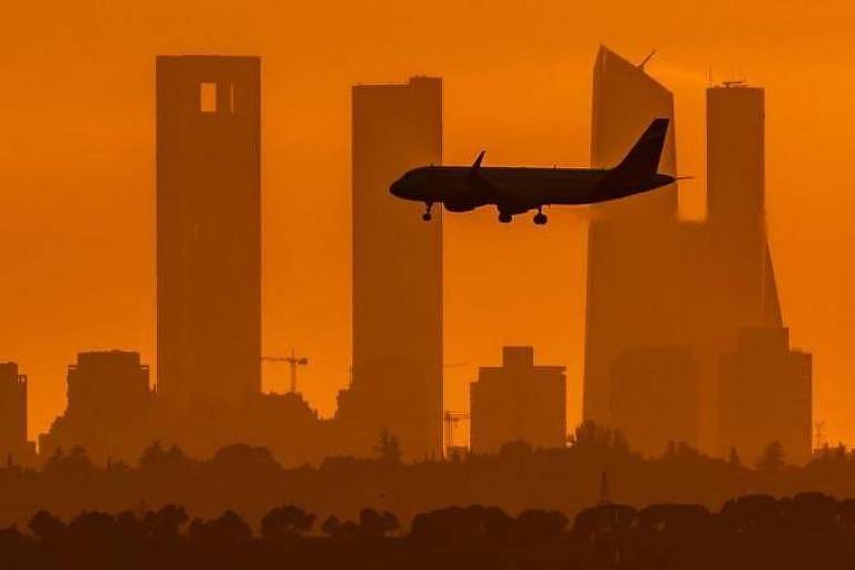 Foto colorida mostra silhueta de avião pousando com silhueta de prédios ao fundo em uma tarde vermelha
