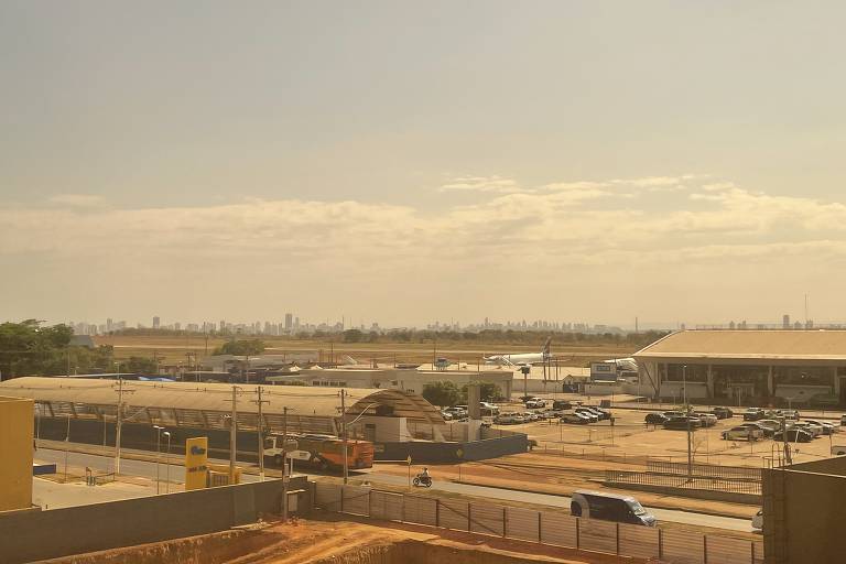 Imagem mostra a estação Aeroporto do VLT de Cuiabá, com o terminal de passageiros e o pátio de aeronaves ao fundo, em Várzea Grande (MT)