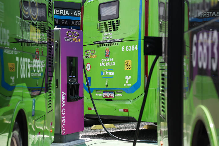 A imagem mostra ônibus verdes estacionados em uma estação de carregamento. Um dos ônibus está conectado a um carregador roxo， que está posicionado em primeiro plano. Os ônibus têm números e logotipos visíveis em suas laterais.