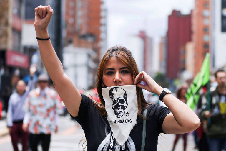 Mulher protesta com braço erguido usando bandana cobrindo a boca; a bandana tem uma caveira e o escrito 'no al fracking'