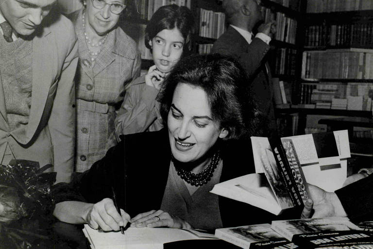 foto em preto e branco de mulher morena autografando livros enquanto sorri