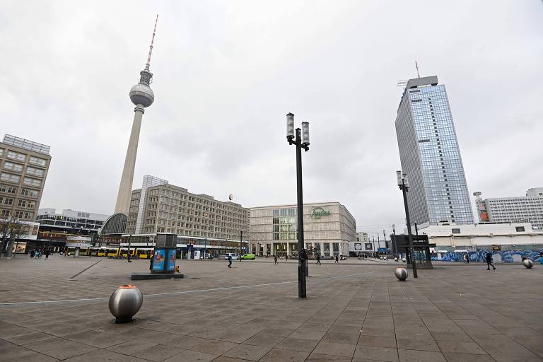 Praça com piso de concreto e, ao fundo, um prédio mais alto à direita, outros mais baixos indo para a esquerda e a torre de TV de Berlim, que é como um obelisco, fina e, no alto, uma esfera. Céu nublado.