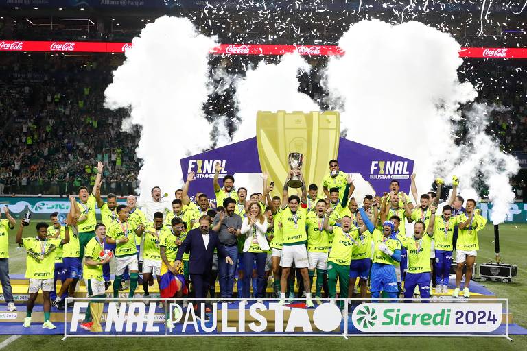 A imagem mostra uma equipe de futebol comemorando a vitória na final do Campeonato Paulista. Os jogadores estão vestidos com uniformes amarelos e verdes， levantando um troféu em meio a fogos de artifício e fumaça. Ao fundo， uma multidão de torcedores celebra. Um banner com a inscrição 039;FINAL PAULISTA039; é visível na parte inferior da imagem.
