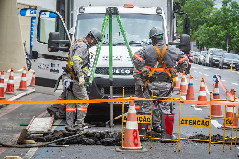 Dois homens com macacão, capacete e faixas luminosas medem buraco em asfalto com caminhão atrás e cones de interdição em volta.