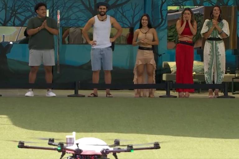 Em foto colorida, seis pessoas observam o pouso de um drone na área externa de uma casa