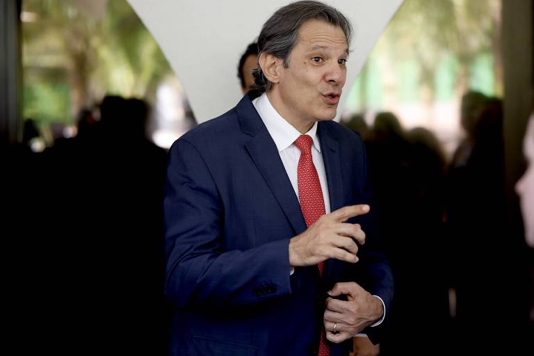 O ministro da Fazenda, Fernando Haddad, ele é um homem branco, tem os cabelos castanhos, usa paletó azul escuro e gravata vermelha e tem o dedo indicador direito apontando para a frente