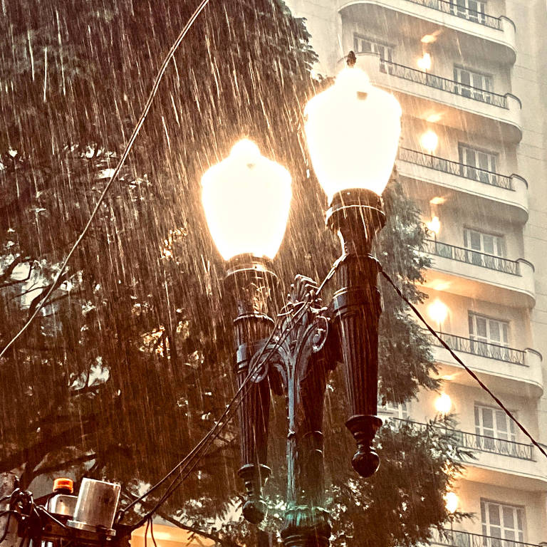 Chuva forte sobre um poste com duas lâmpadas em uma calçada, com um prédio ao fundo