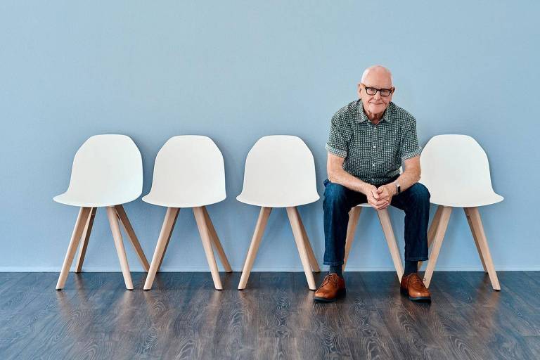 Homem de cabelo grisalho está sentado em uma cadeira branca num sala de espera. A parede é azul.