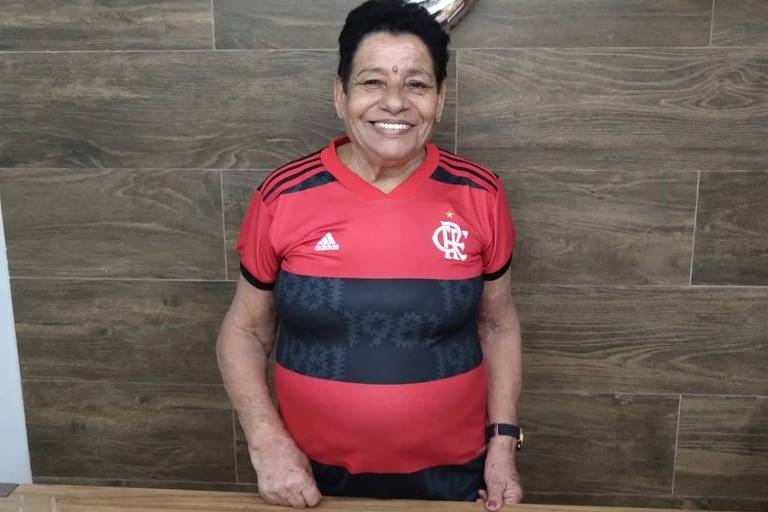olivia é idosa, tem cabelos pretos curtos, sorri para foto, usa camisa do Flamengo, time de futebol