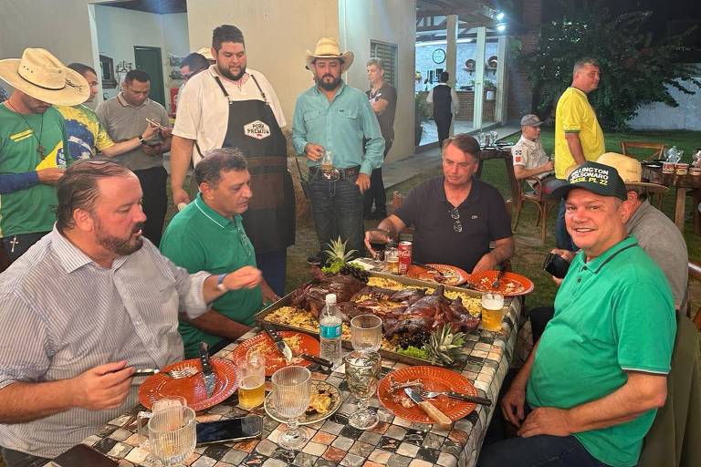 Foto mostra Bolsonaro sentado em uma mesa onde é servido um churrasco de porco. 