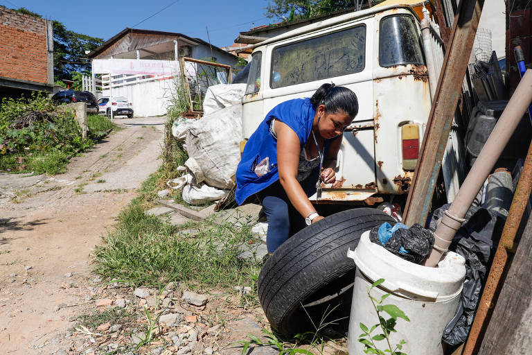 Mulher vigilante de saúde com colete azul mexe em pneu em busca de água parada em um lugar com entulho acumulado durante mutirão