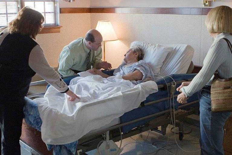 Fotografia colorida mostra uma mulher branca e idosa em um leito de hospital cercada por outras duas mulheres e um homem, todos brancos