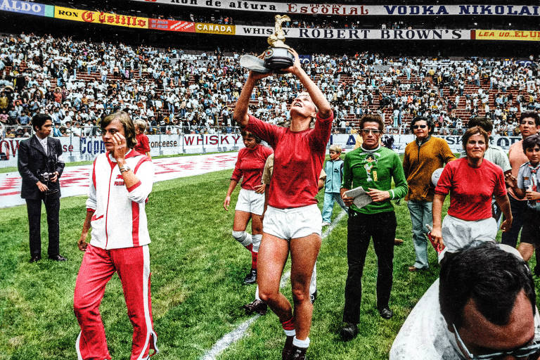 Mulher branca e loira veste shorts branco e camisa vermelha com manga longa. Ela segura um troféu dourado enquanto anda em um gramado verde. Ao fundo, o público lota as arquibancadas do estádio de futebol.