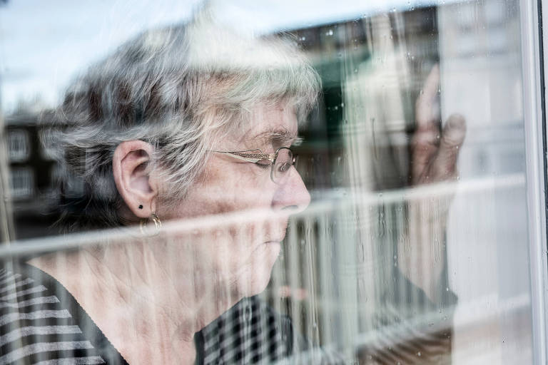 Uma senhora idosa olha pensativamente através de uma janela, com seu reflexo capturado no vidro, sugerindo uma introspecção profunda ou uma lembrança distante. 