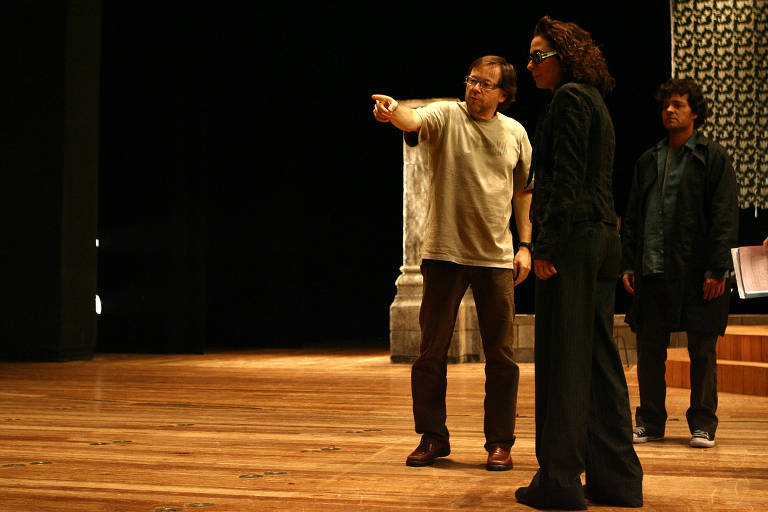 Três pessoas, dois homens e uma mulher, estão no palco do Theatro Municipal. À esquerda, o diretor Fernando Meirelles tem o braço estendido apontando para algo. Os dois atores observam