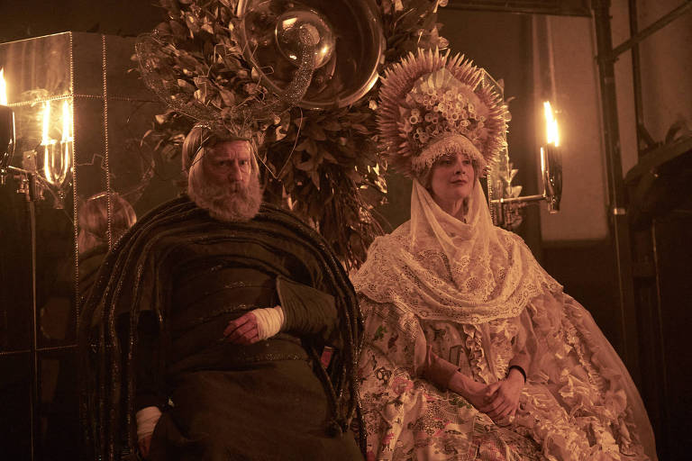 Um homem e uma mulher sentados em tronos vestidos com roupas suntuosas e coroas enormes 