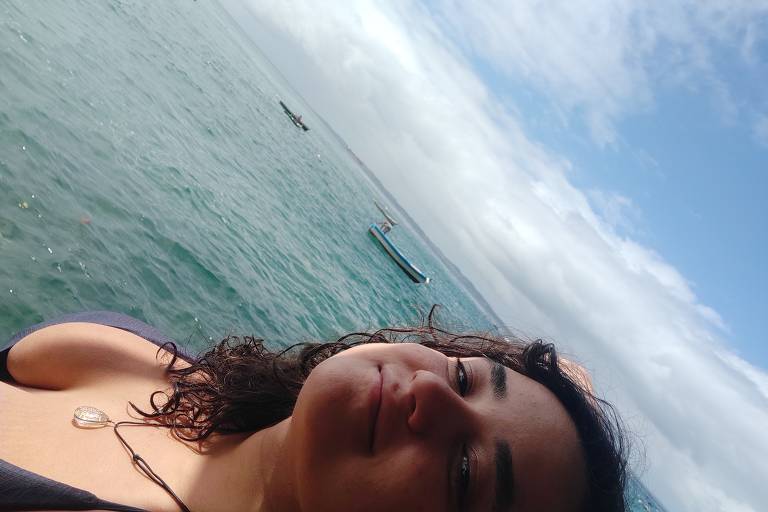 Mulher branca de cabelos escuros usa um biquíni preto e tira uma selfie de frente ao mar